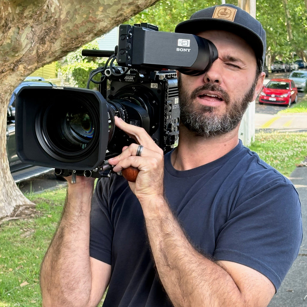 Jono Drew camera operator holding a Sony camera with Wooden Camera handle outdoors with trees and cars in the background
