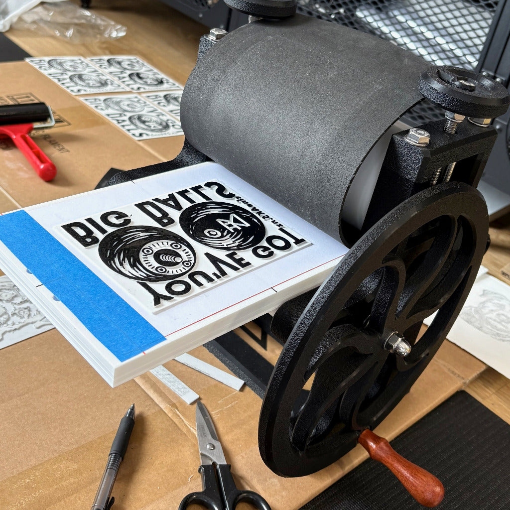 Printing press with a design on a sheet of paper, surrounded by tools on a wooden surface.