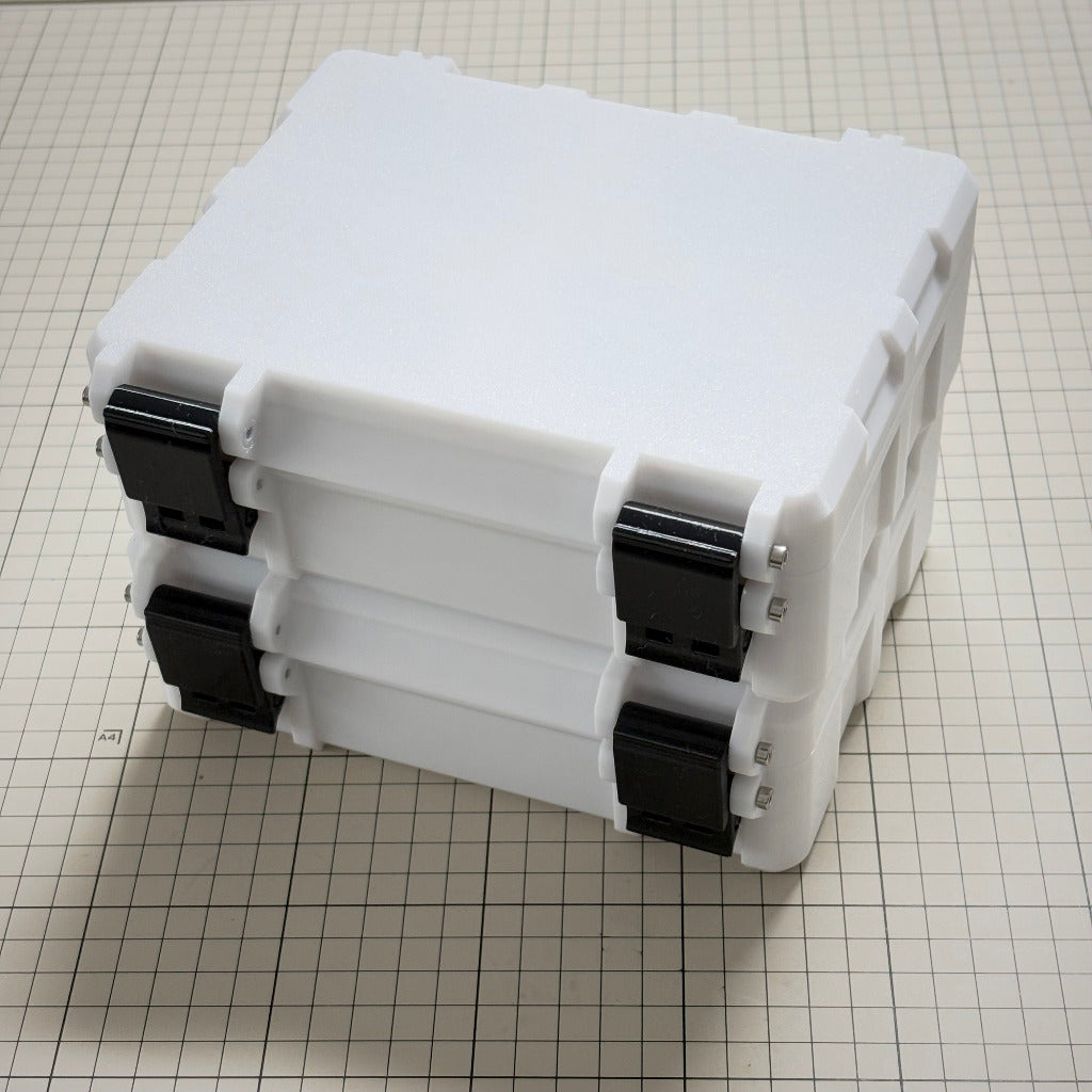 Two stacked white plastic storage boxes with black latches, photographed on a beige grid cutting mat.