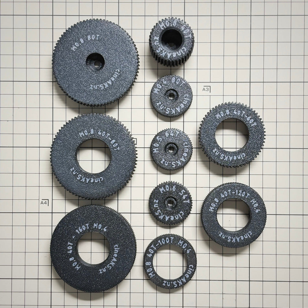A set of cineAKS cforce Starter Kit M0.8, M0.6, M0.5 and M0.4 / 64p lens gears, arranged on a white grid background. Each wheel has white text printed on it including the brand 'cineAKS.nz' and size specifications.