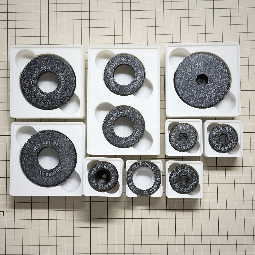 A set of cineAKS cforce Starter Kit with 10 direct-fit and push-on cinema lens gears arranged in white gridfinity trays on a beige grid background. The set includes several larger and smaller pieces, with some showing white stamped text "cineAKS.nz", and sizes.