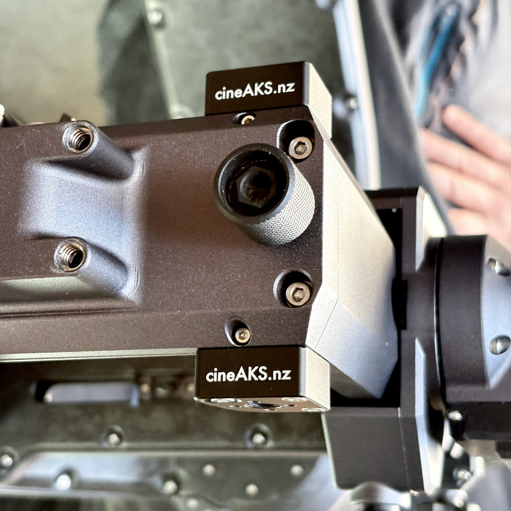 Close-up of a CineAKS camera accessory MotoCrane TitanLT replacement corner bracket with ARRI locating 3-8-16 thread with two visible CineAKS.netz labels, showing the dark metallic body, mounting hardware, and part of a hand in the blurred background.