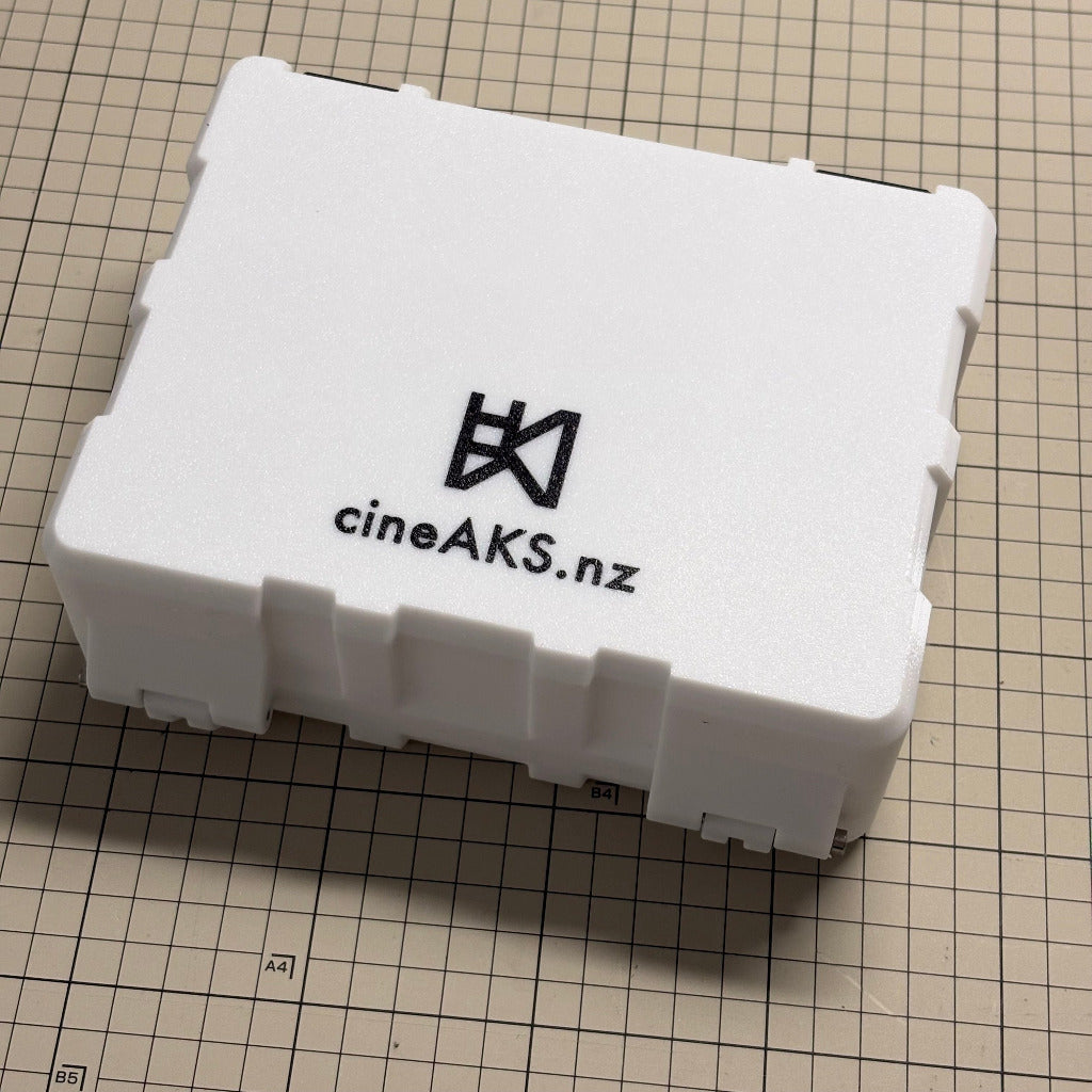 A gridfinity compatible white rectangular hard case with a black printed CineAKS logo and 'cineAKS.nz' on the lid, placed on a beige gridded cutting mat.